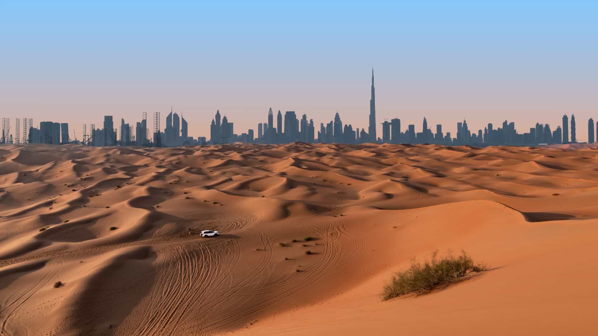 Dubai skyline with the Burj Khalifa in the distance behind sunlit desert sand dunes with tire tracks and a small vehicle.