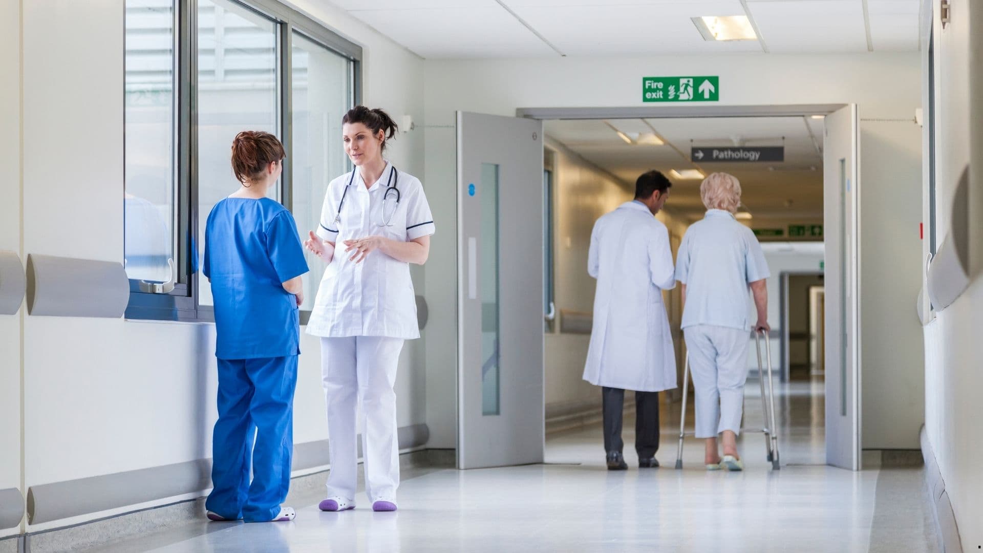 Nurse and healthcare worker talking in a hospital corridor as a doctor and patient walk toward the pathology department.