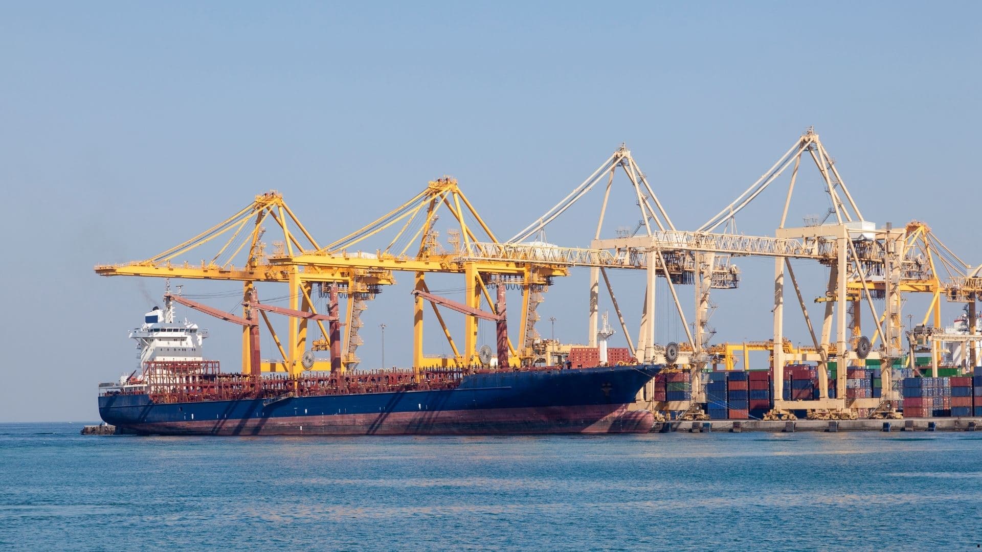 Container ship berthed at a port with gantry cranes loading cargo, representing trade and logistics operations in scope of emissions reporting.