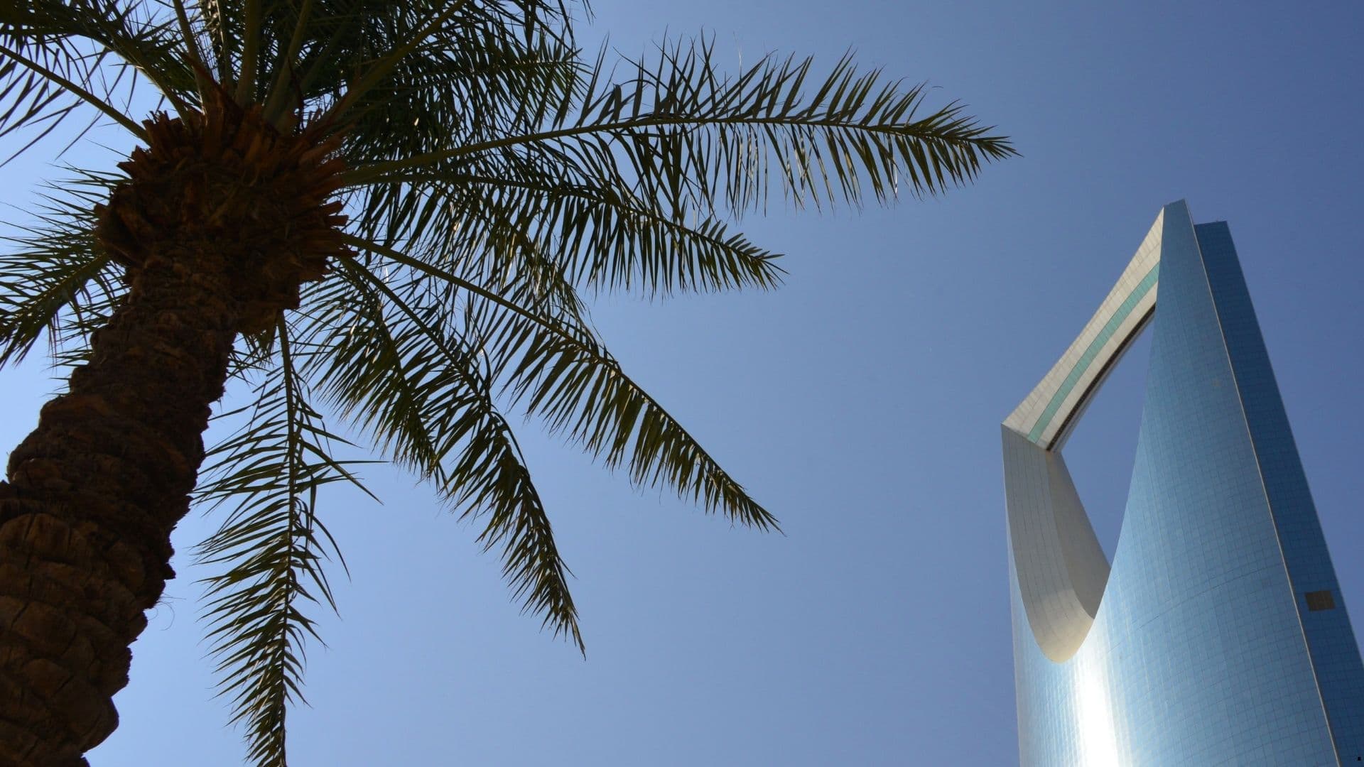 Kingdom Centre tower in Riyadh, Saudi Arabia, seen from below against a clear blue sky with a palm tree in the foreground.