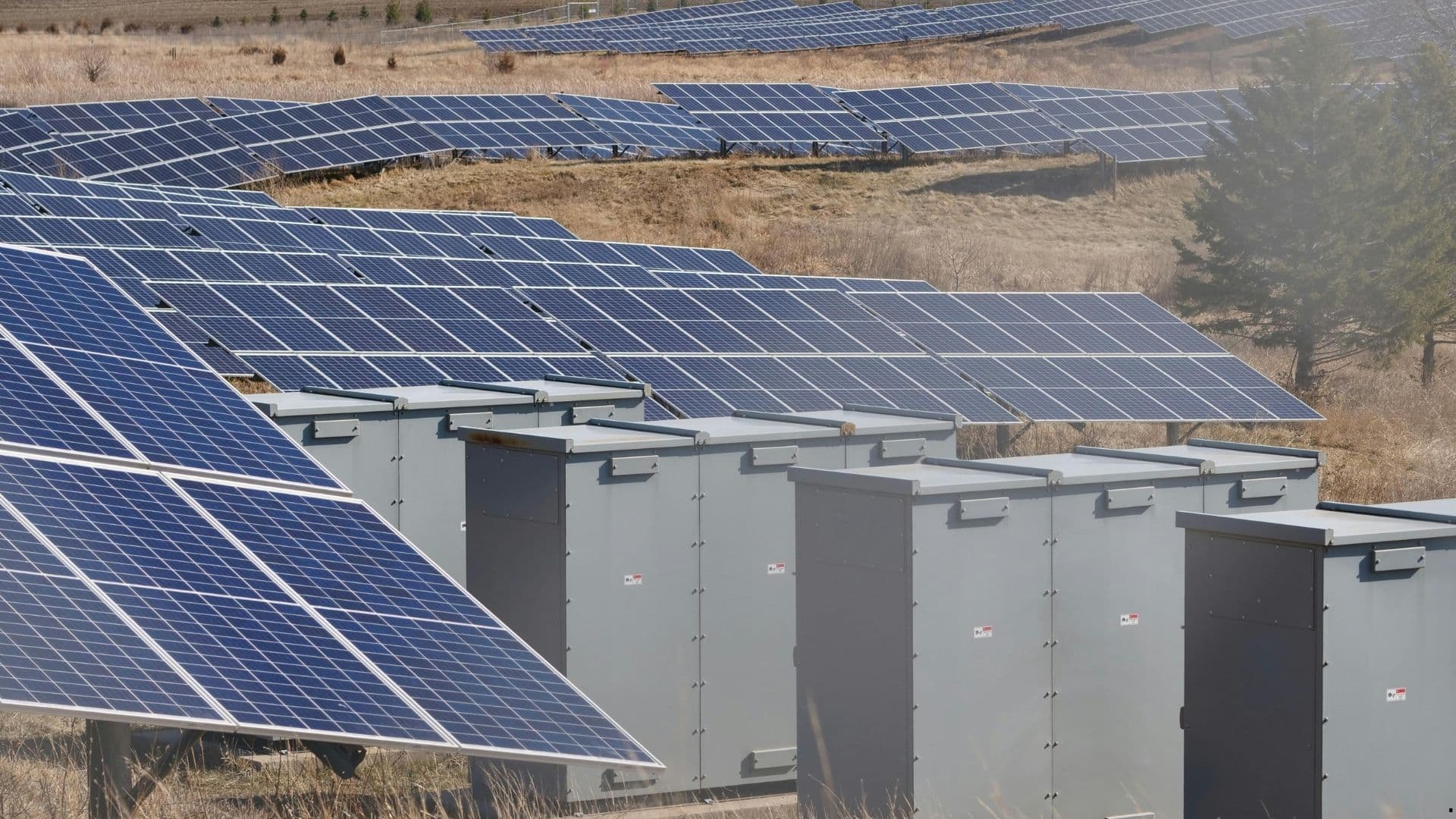 Rows of solar panels beside grey battery and power-conversion units at a solar-plus-storage site.